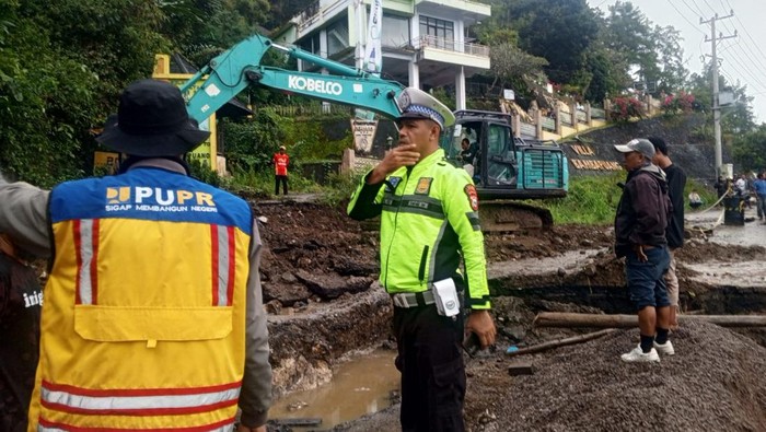 Seorang polisi lalu lintas dan petugas PUPR mengawasi proses perbaikan jalan amblas di Jalan Poros Enrekang-Toraja, dengan alat berat ekskavator bekerja di latar belakang.
